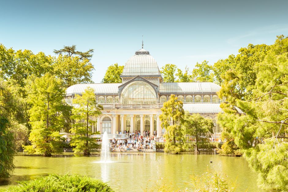 A large glass greenhouse with a domed roof and intricate metal framework is visible, situated behind a lush pond surrounded by tall green trees and shrubs. In front of the greenhouse, a group of people can be seen standing on a paved area near the entrance, which features classical columns and ornate architectural details. The scene is set during daytime with clear blue skies and bright natural lighting, highlighting the vibrant foliage and reflections on the calm water of the pond. This image captures the exterior of a conservatory or botanical building, possibly within Crystal Palace Park, illustrating the environment where house removals and furniture transport services by Man and Van Crystal Palace might operate, especially when planning access and timing for home relocation projects.