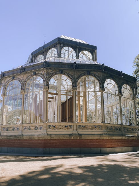 A large, ornate glass conservatory with intricate metal framing and decorative details, situated outdoors under a clear blue sky. The structure includes multiple arched windows and a domed top section, with visible reflections of the sky and surroundings on the glass panels. The area around the conservatory appears to be part of a park or landscaped setting, which aligns with the context of a house removals or relocation service. Man and Van Crystal Palace may utilize such landmarks during the logistics of home relocation or furniture transport, as part of their tips on access and timing for household moves in the Crystal Palace area.
