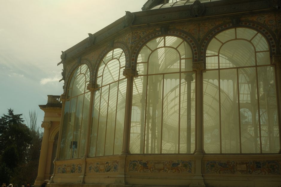 Photograph of the ornate glass and metal framework of a historic greenhouse or conservatory, featuring large arched windows with intricate metal detailing and decorative supports. The structure is ill