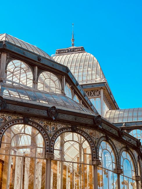 A detailed exterior view of a historic glasshouse with ornate metal framing and multiple curved glass panels, topped with a domed central structure and a decorative spire, set against a bright blue sk