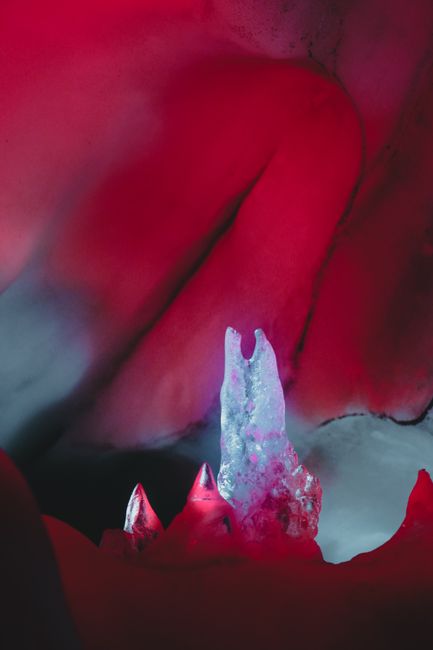 The image shows the inside of a cave or crevice illuminated with red lighting, highlighting jagged rock formations with sharp, pointed peaks. In the foreground, a large ice formation with a translucent, bluish-purple hue stands prominently among the rocks, suggesting a natural formation or decorative element. Surrounding the ice, there are smaller ice or mineral deposits with similar coloration. The environment appears confined, with the rocks and ice forming a compact, intricate landscape. This setting could be part of a natural show cave or an artistic installation designed to resemble a frozen cavern. The lighting emphasizes the textures and shapes of the rocks and ice, providing a vivid visual experience that might be relevant for illustrating home relocation or moving logistics involving fragile or decorated items like ice sculptures or aesthetic interior features. Man and Van Crystal Palace, specialists in removals, might use such imagery to depict the careful handling and transport of delicate or distinctive objects during a furniture or home relocation process.