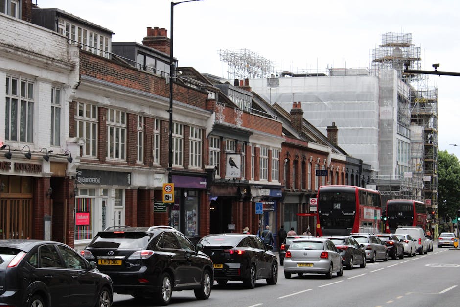 A busy street scene in Westow Hill, Crystal Palace, showing a row of traditional Victorian-style terraced shops with large display windows and brick facades. In front of the shops, several vehicles including black, white, and grey cars are parked along the roadside, with some traffic congestion visible as cars wait in traffic. Two red double-decker buses are moving along the street, with one approaching and another further behind. The pavement area next to the shops features pedestrians walking, while some individuals are engaged in loading or unloading items. Behind the shops, a building under construction is covered in scaffolding and white sheeting, indicating ongoing renovation or development. The scene is well-lit with natural daylight, capturing the typical urban environment suitable for house removals or furniture transport services, as coordinated by companies like Man and Van Crystal Palace, aiming to facilitate home relocations in tightly built streets like Westow Hill.