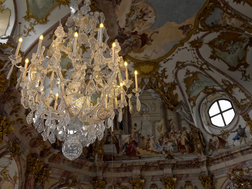 Close-up view of an ornate crystal chandelier hanging from the ceiling of a historic room, with multiple lit candle-shaped bulbs and intricate glass designs. The ceiling features elaborate gold and white decorative mouldings and a painted fresco, with a round window providing natural light. The chandelier is prominently centered, with surrounding elements including detailed wall decorations and partial view of the ceiling artwork. This setting reveals a luxurious interior aligned with traditional European interiors, relevant to home or building removals involving delicate fixtures, and may be associated with detailed packing and careful transportation by Man and Van Crystal Palace during furniture transport or relocation processes.